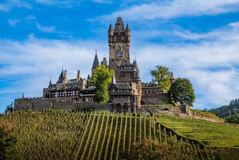 Cochem, Germany A breathtaking view of Reichsburg Castle, set atop a lush hill, surrounded by rows of vineyards in the scenic Moselle Valley.