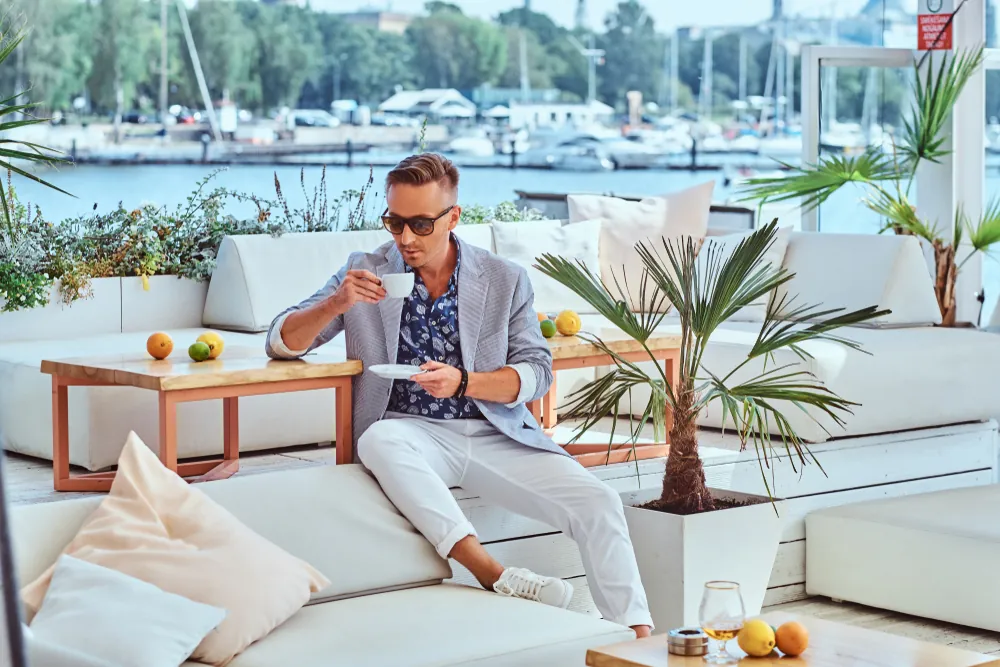 Fashionable successful man with stylish hair dressed in modern elegant clothes holds a cup of coffee while sitting on a sofa at outdoor cafe against the background of the city wharf.